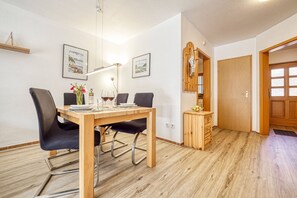 Un coin repas avec une table en bois, des chaises noires et un luminaire suspendu. On y trouve des tableaux encadrés au mur, un meuble en bois et une porte avec des panneaux de verre.