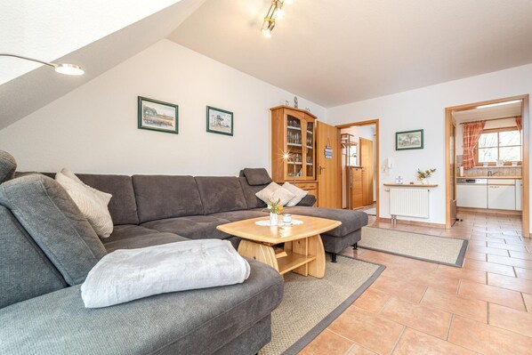 A cozy living room with a grey sectional sofa, a wooden coffee table, and a kitchen visible through an open doorway. - Comfortable vacation apartment in a vacation park with balcony and pool in Zingst - (Zingst)