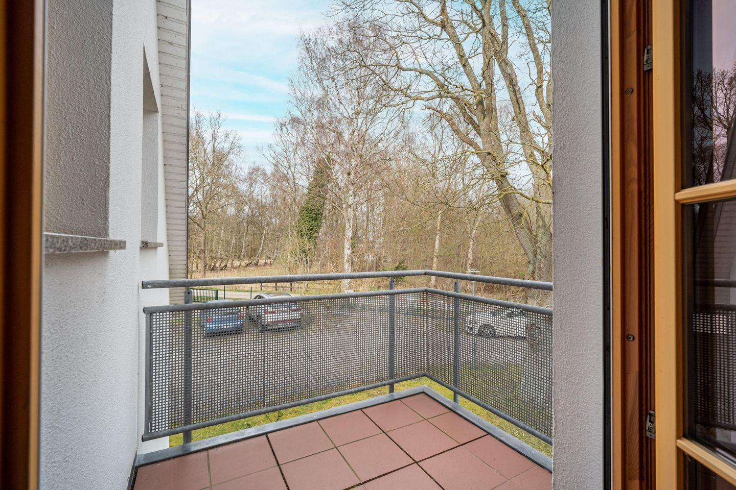 Ein Balkon mit Metallgeländer, Fliesenboden und Blick auf Bäume sowie ein geparktes Auto.
