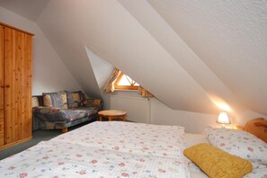 Um quarto de sótão acolhedor, com teto inclinado, guarda-roupa de madeira, sofá com almofadas estampadas, mesa redonda de madeira e cama com roupa de cama florida.