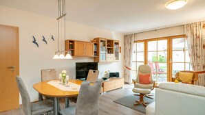 Ein modernes Wohnzimmer mit Essbereich, Holzkredenzen, einem Fernseher und einem großen Fenster mit Vorhängen.