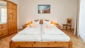 Una cama doble con ropa de cama blanca, dos mesitas de noche con lámparas, un armario de madera, una silla y un cuadro en la pared.