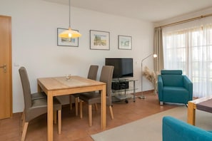 Un salon comprenant une table à manger en bois, deux chaises, un téléviseur posé sur un meuble en verre, un fauteuil bleu et une fenêtre ornée de rideaux légers.