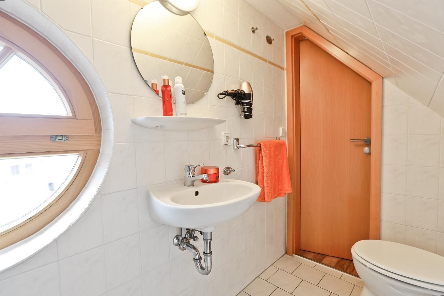 Um banheiro com uma janela redonda, pia com espelho acima, secador de cabelo e vaso sanitário.