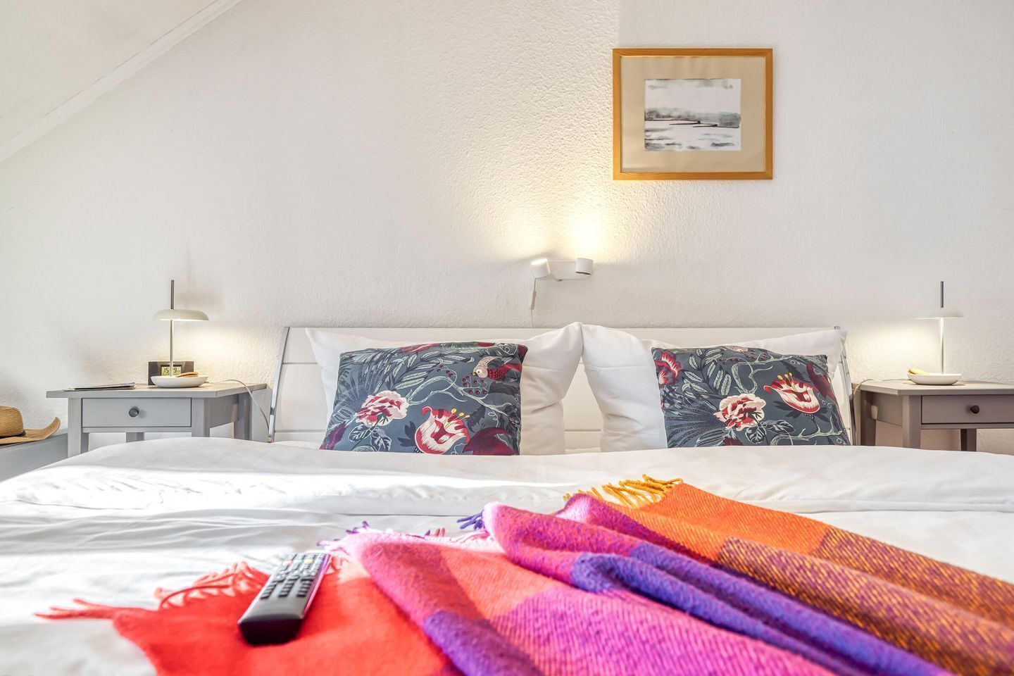 A hotel room with a bed, two nightstands, a lamp, a remote control, and a framed picture on the wall.