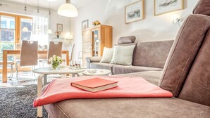 A modern living room with a brown sofa, a dining table with chairs, and a kitchen area in the background. - Comfortable terraced house in a vacation park with terrace and garden in Zingst - (Zingst)