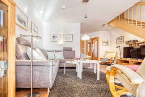 Uma sala de estar com um sofá cinzento, uma mesa de centro branca, uma cadeira de madeira e uma televisão.