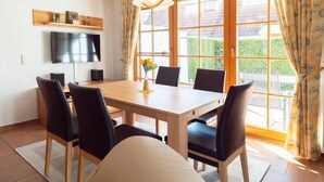 A dining area with a table, chairs, a TV, and a window with curtains. - Terraced house in Zingst with terrace, garden view and heated indoor pool - (Zingst)