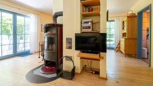 A modern living room with a wood-burning stove, a flat-screen TV, and a bookshelf. - Comfortable semi-detached house with sauna, garden and terrace in the Zingst vacation park (Zingst)