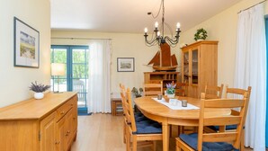 A dining area with a wooden table and chairs, a chandelier, and a view of the outdoors through a door with curtains. - Comfortable semi-detached house with sauna, garden and terrace in the Zingst vacation park (Zingst)