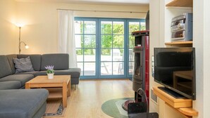 A living room with a sofa, coffee table, and a TV. There is a large window with curtains, a fireplace, and a bookshelf. - Comfortable semi-detached house with sauna, garden and terrace in the Zingst vacation park (Zingst)