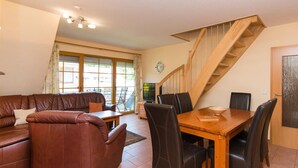 Ein Wohnzimmer mit braunem Ledersofa, einem Holzesstisch mit Stühlen, einer Treppe und einem großen Fenster mit Vorhängen.