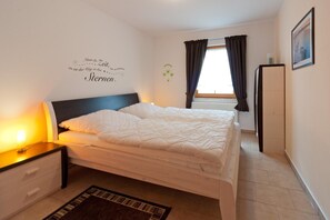 A bedroom with a bed, a nightstand, a wall decoration, a window with curtains, and a radiator.