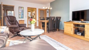 A modern living room with a sofa, a dining table with chairs, a TV, and a wooden cabinet with electronic devices. - Spacious Möwennest vacation apartment with terrace, garden view and pool in Zingst (Zingst)