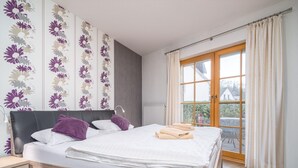 A neatly made bed with white linens and purple pillows, a wooden nightstand, and a large window with curtains. - Spacious Möwennest vacation apartment with terrace, garden view and pool in Zingst (Zingst)