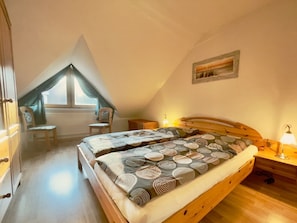 Une chambre avec un plafond en pente, un lit avec une literie à motifs, une chaise, une table de chevet avec une lampe et une fenêtre avec des rideaux.