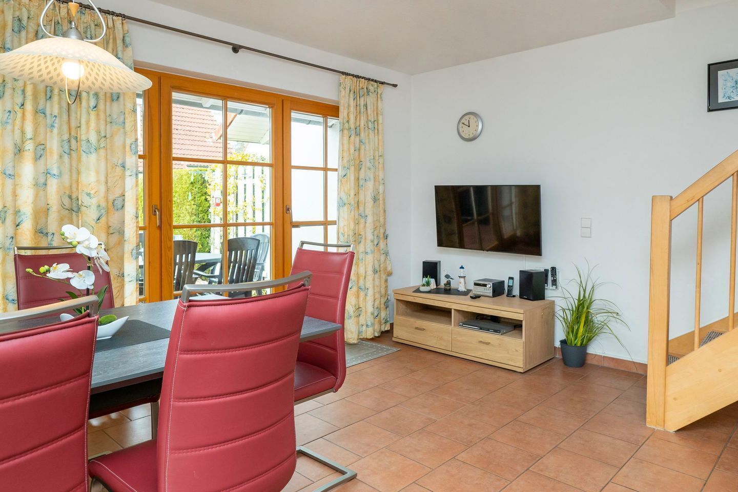 A dining area with red chairs and a table, a TV on a wooden stand, and a clock on the wall.