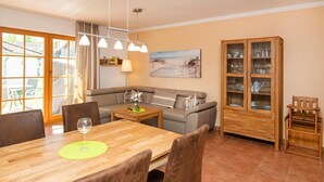 A dining area with a wooden table, chairs, and a glass of wine. There is a sofa and a cabinet with glass doors in the background. - Comfortable vacation apartment with terrace, garden view and heated pool in Zingst (Zingst)