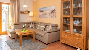 A living room with a sofa, a coffee table, a cabinet with glass doors, a painting on the wall, and a window with curtains. - Comfortable vacation apartment with terrace, garden view and heated pool in Zingst (Zingst)