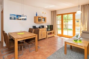 Una sala de estar acogedora con una mesa de comedor de madera, un sofá y un mueble para la televisión. Hay dos ventanas con cortinas, un aparador con puertas de cristal y una pequeña mesa con una lámpara.