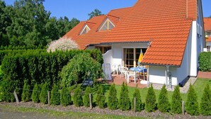 A house with a red-tiled roof, a covered patio with outdoor furniture, and a well-maintained garden. - Comfortable terraced house with terrace, garden and pool in the Freesenbruch vacation park (Zingst)