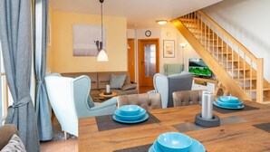 A modern living room with a dining table set for four, a sofa, a TV, and a staircase. - Comfortable terraced house with terrace and garden in the Freesenbruch vacation park - (Zingst)
