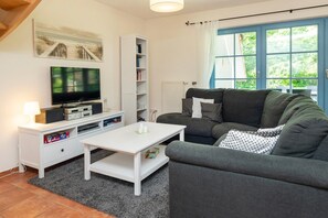 A living room with a television, white entertainment unit, grey sofa, and a window with curtains. - Cozy townhouse with a terrace, a view of the garden, and a pool in Zingst - (Zingst)