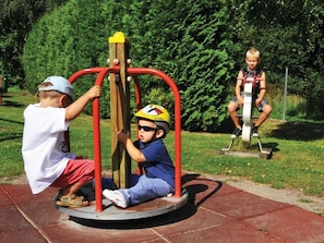 A playground with a rotating play structure, a grassy area, and a metal fence in the background. - Cozy townhouse with a terrace, a view of the garden, and a pool in Zingst - (Zingst)