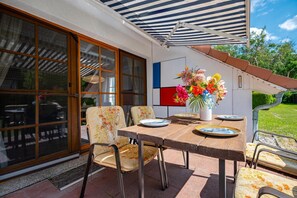 An outdoor patio with a table set for four, surrounded by chairs with floral upholstery, a large window with blinds, and a colorful flower arrangement. - Cozy townhouse with a terrace in the Freesenbruch vacation park near the sandy beach (Zingst)