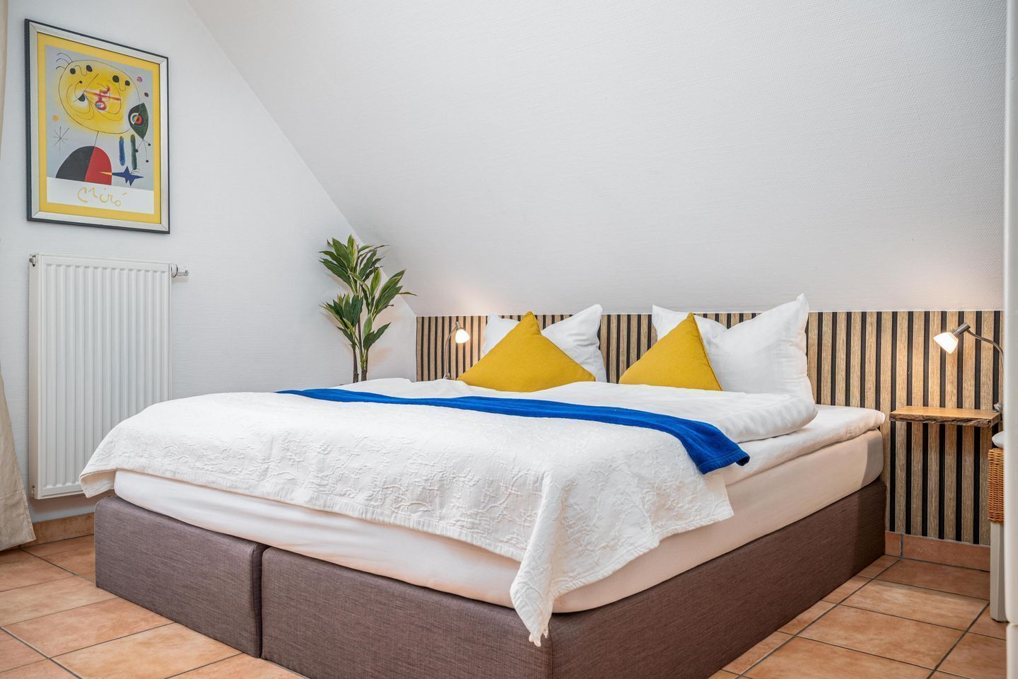 Une chambre à coucher avec un lit, une table de chevet, une lampe, un radiateur et un tableau encadré au mur.
