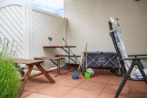 A patio with a wooden table, a folding table, a wheelbarrow, and a blue storage unit with a logo. - Semi-detached house lighthouse in vacation park with garden, terrace and pool - (Zingst)