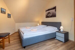 Un dormitorio moderno con cama gris, ropa de cama blanca, dos mesitas de noche con lámparas, una silla de madera y un cuadro de un barco en el agua.