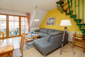 Una sala de estar acogedora con un sofá gris, una mesa de comedor de madera y sillas, una escalera con barandillas verdes y una ventana con cortinas.