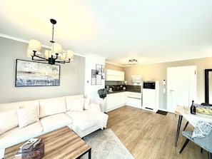 Ein modernes Wohnzimmer mit Sofa, Couchtisch und Essbereich mit Tisch und Stühlen. Im Hintergrund ist eine Küche mit weißen Schränken und Geräten erkennbar.