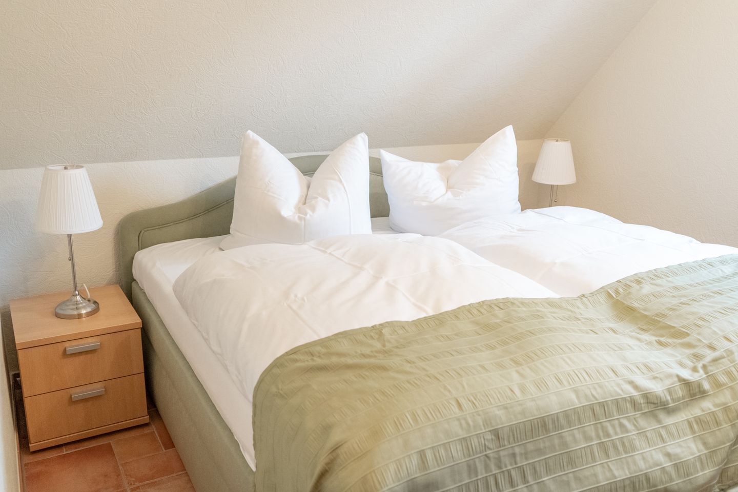 Een netjes opgemaakt bed met witte lakens en een groen gestreept dekbed, een bedlamp en een houten nachtkastje met een lade.