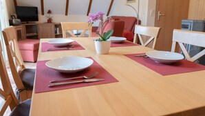 A dining area with a wooden table set for four, surrounded by chairs. There is a potted plant on the table, and a television is visible in the background. - Haus Sabine - Upper floor - Bright, large upper floor apartment in a quiet location! (Zingst)