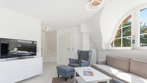 Un salon moderne avec un téléviseur à écran plat, un fauteuil gris, un canapé blanc et une petite table sur laquelle repose un magazine.