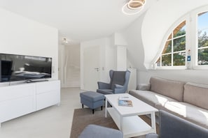 A modern living room with a flat-screen TV, a grey armchair, a white sofa, and a small table with a magazine.