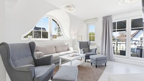 Un salon moderne comprenant un canapé, des fauteuils, une table basse et une table à manger. De grandes fenêtres, ornées de rideaux, offrent une vue sur l’extérieur, et le plafond présente un design incurvé original.