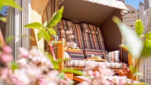 Une chaise en osier avec des coussins à rayures et une table en fibres tressées, placées sur une véranda d’où l’on peut admirer un bâtiment.