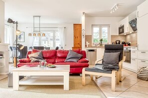 Una sala moderna con un sofá de cuero rojo, una mesa de centro de madera y un sillón gris. Se ve una cocina en el fondo, con gabinetes blancos y un horno empotrado.