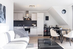 Et moderne køkken med hvide skabe, en sort bordplade og rustfrie stålapparater. Der er et spiseområde med et rundt bord og stole, samt en stue med en sofa og et sofabord.