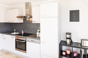 Une cuisine moderne avec des meubles blancs, une hotte en acier inoxydable, un lave-vaisselle et une machine à café posés sur un plan de travail noir.