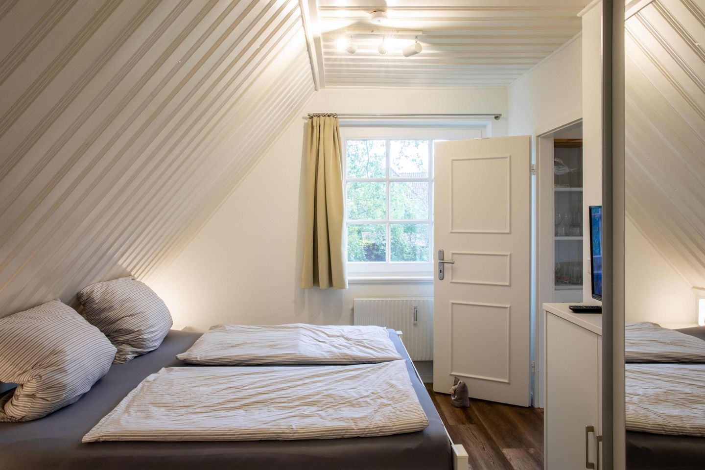 A bedroom with a sloped ceiling, a window with curtains, a bed with a blanket, and a wardrobe.