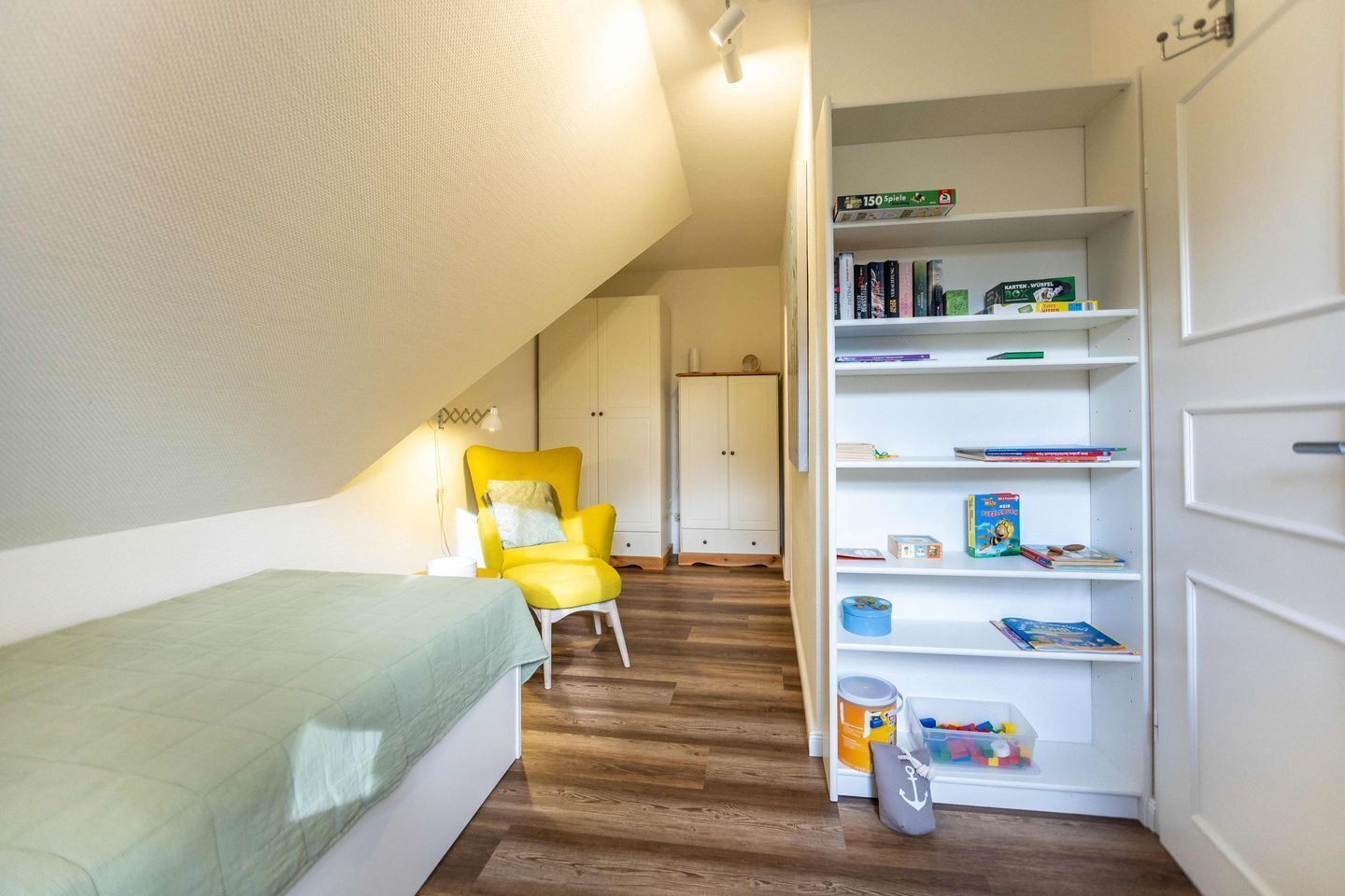 A small, cozy room with a single bed, a yellow chair, and a bookshelf filled with books and toys.