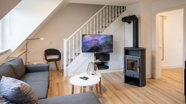 A modern living room with a grey sofa, a flat-screen TV, a black wood-burning stove, and a staircase with white railings. - Vacation apartment Wrixumer Marsch - Located directly on the marsh (Wrixum)