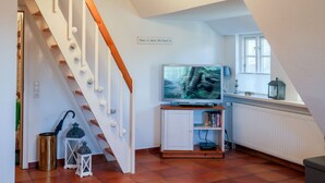 A room with a white staircase, a television on a stand, a bookshelf, a radiator, and a window with a view of trees. - Ferienwohnung Britta (Wyk auf Föhr)