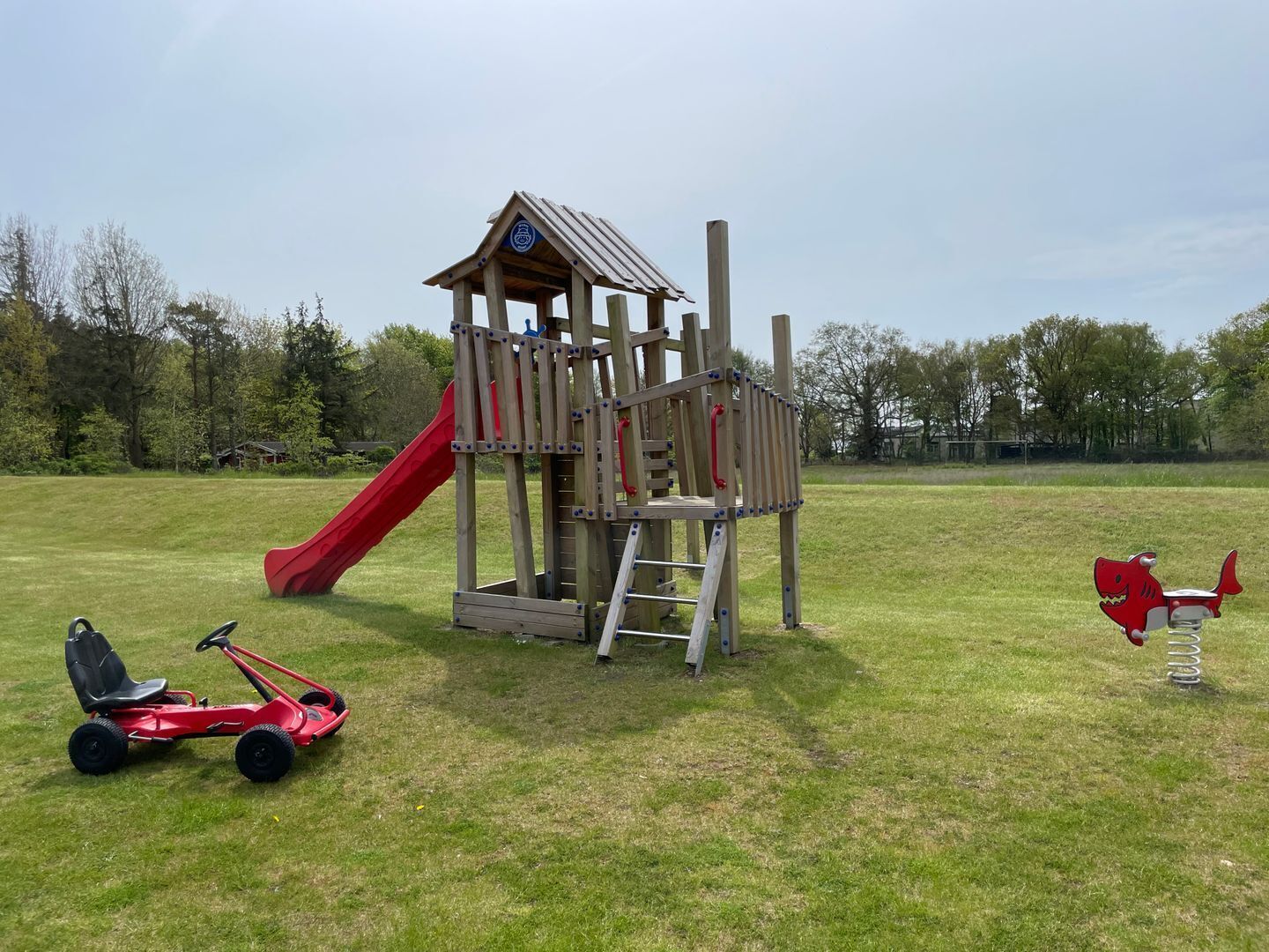 Eine hölzerne Spielkonstruktion mit Rutsche und Kletterelementen, ein rotes Dreirad und ein rotes Spielzeugauto.