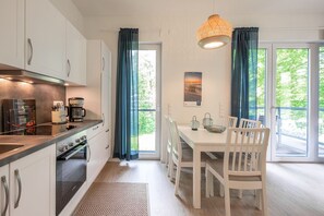 Eine moderne Küche mit weißen Schränken, einem eingebauten Backofen und einer Kaffeemaschine. Ein Esstisch mit weißen Stühlen bietet Blick auf grüne Natur durch die Fenster.
