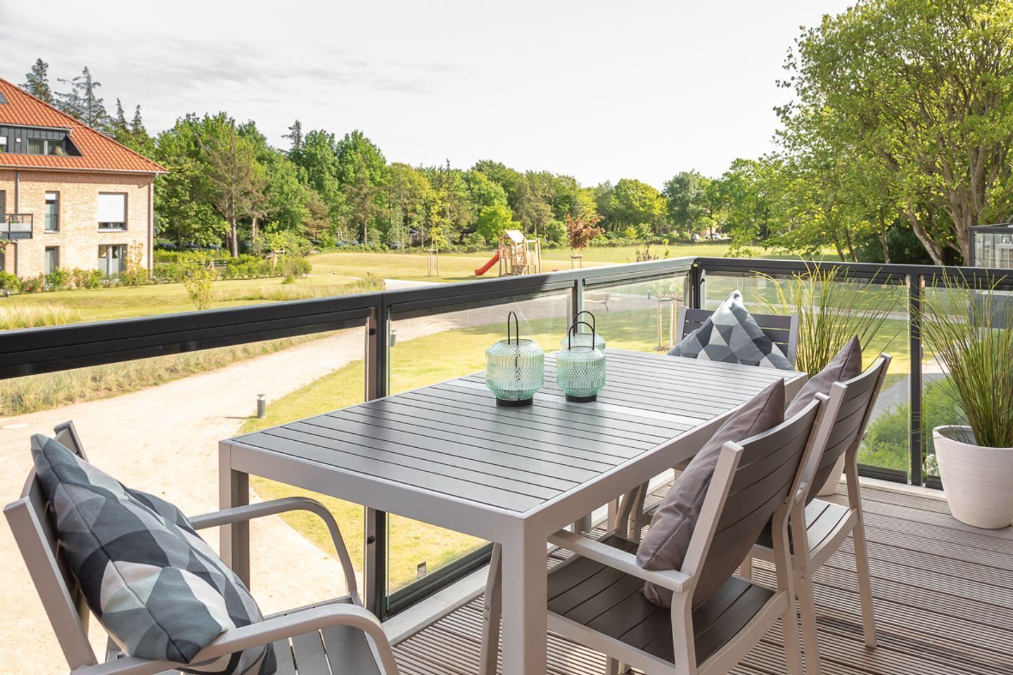 A balcony with a table and chairs, a potted plant, and a view of a playground and trees.